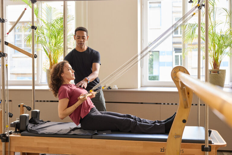 Trainer correcting the torso posture of a female client sitting on the Reformer.