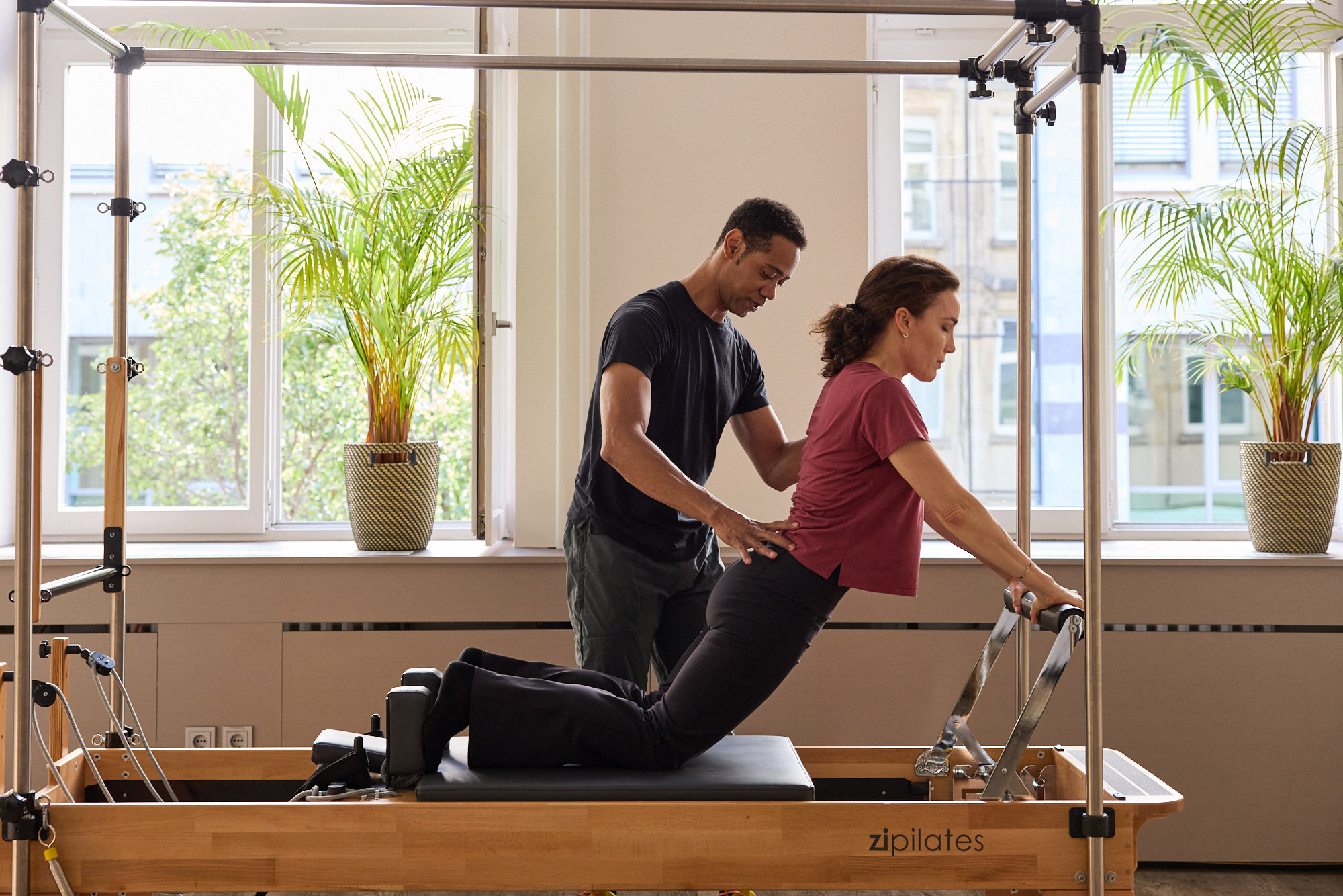 Trainer unterstützt eine Klientin bei einer kniend ausgeführten Übung am Pilates Reformer Cadillac-Gerät.