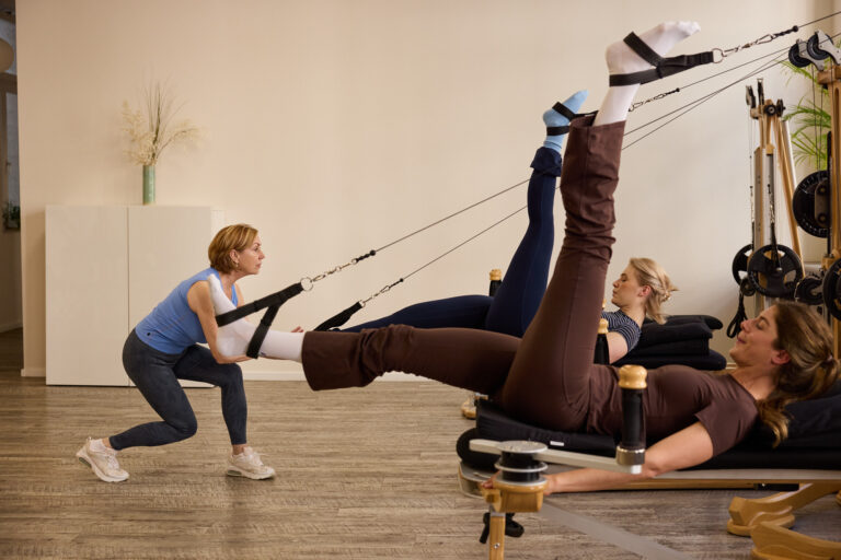 A trainer guides a client through a leg extension movement using strap resistance on a Gyrotonic Tower.
