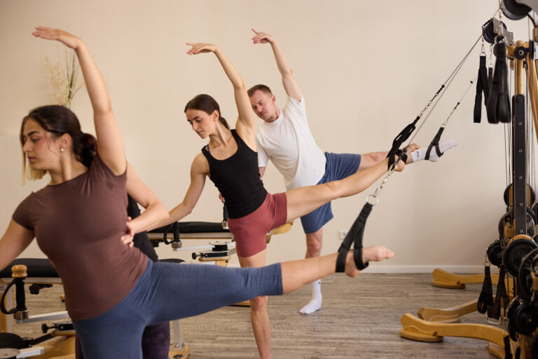 Three female dancers lift their legs, supported by the Gyrotonic Pulley Tower, while extending their arms overhead and stretching to the side.