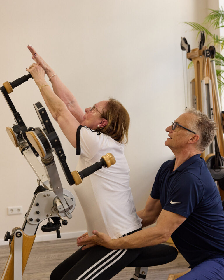 Trainer unterstützt eine Klientin bei einer Streckübung am Gyrotoner – Rehabilitation.
