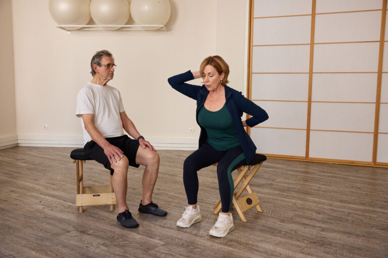 A trainer demonstrates a seated exercise with a hand on the head, observed by a client.