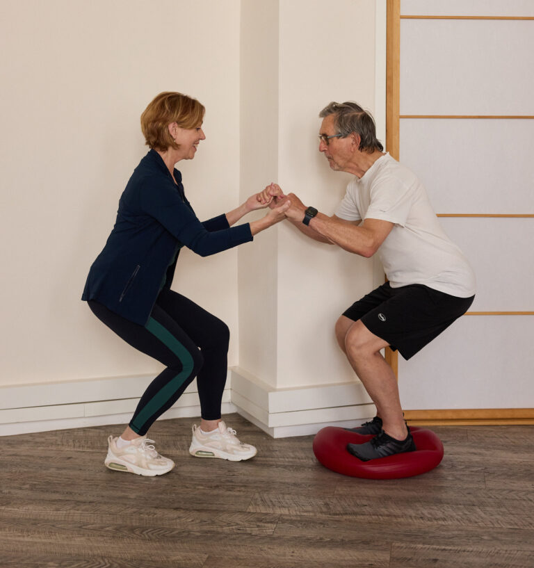 A trainer assists a client with a balance exercise on a cushion.