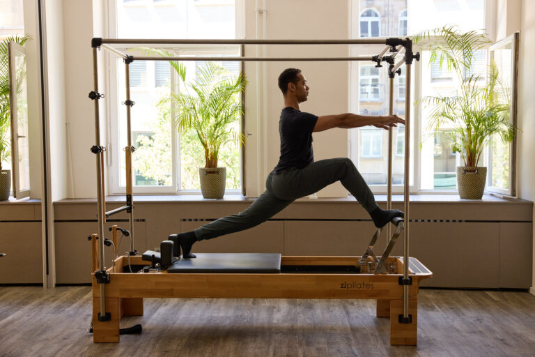 A man is exercising in a lunge position on a Pilates Reformer Cadillac.