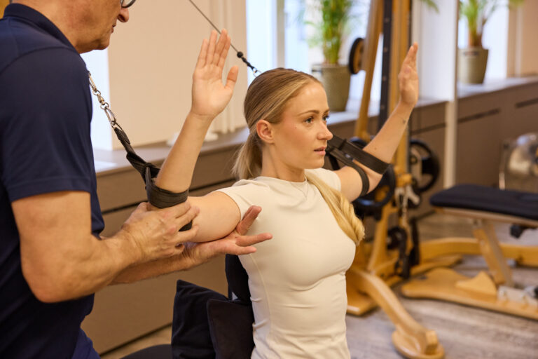 Trainer assisting a client on the Gyrotonic machine with arms raised.