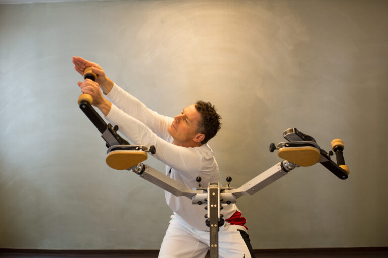 Man exercising with outstretched arms on a Gyrotonic machine against a neutral background.
