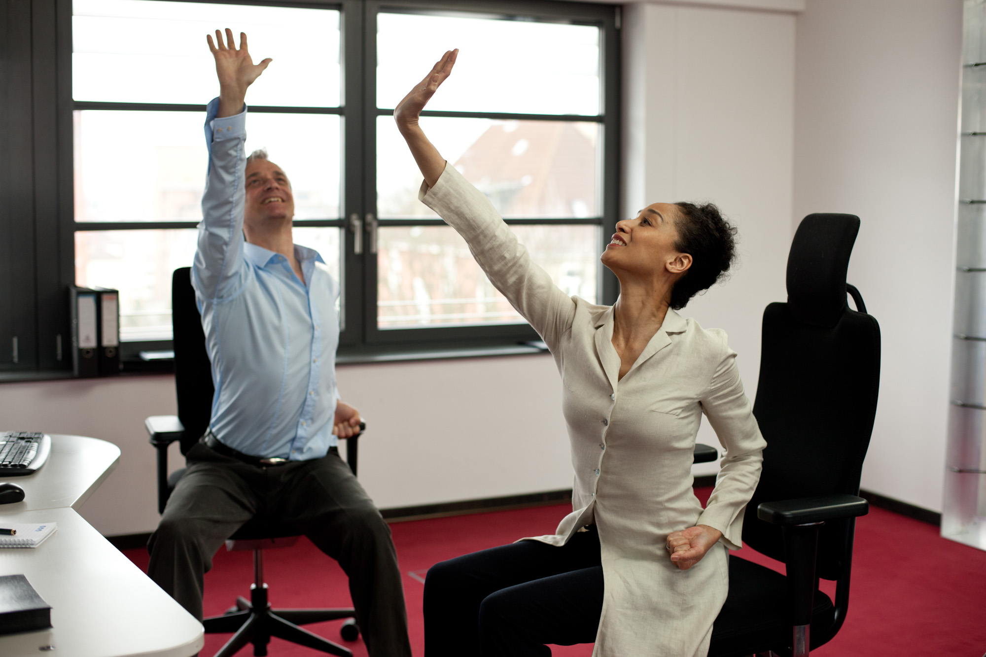 Two colleagues are stretching on office chairs with their arms raised.
