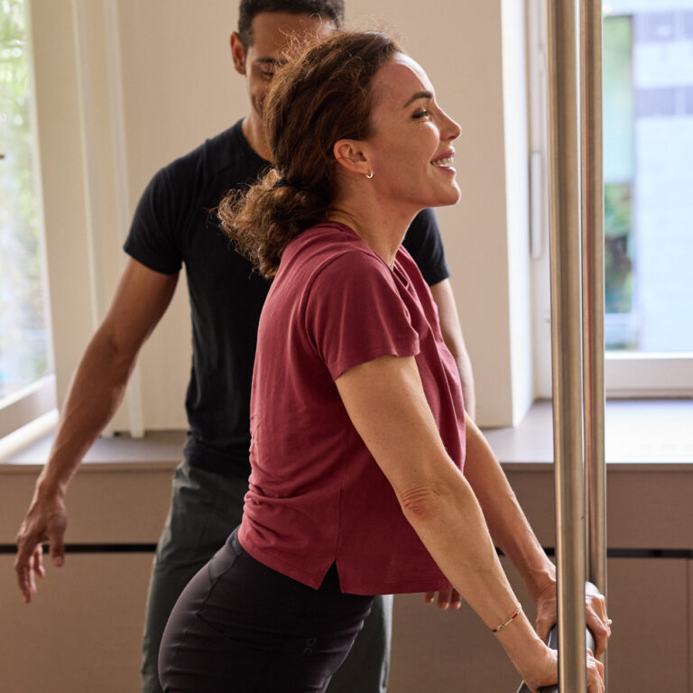 Trainer betreut lächelnde Klientin kniend am Pilates Reformer Cadillac